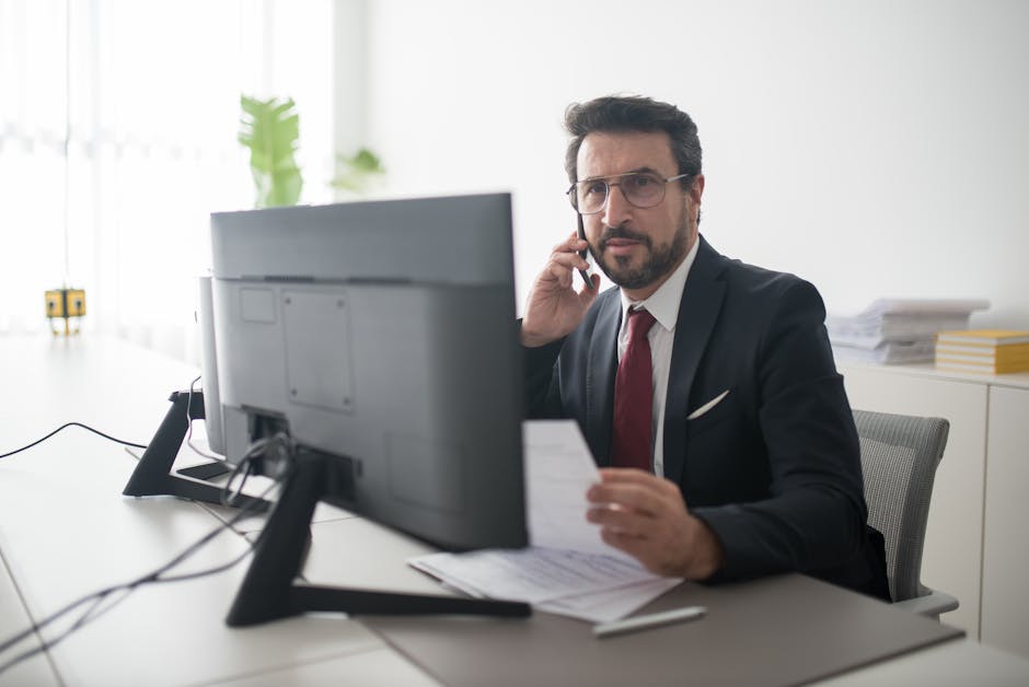 Confident businessman in a black suit making a phone call while reviewing documents in a modern office setting.