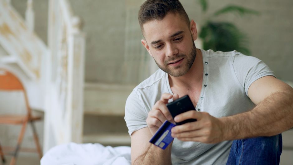 Young man using smartphone and credit card for online shopping indoors.