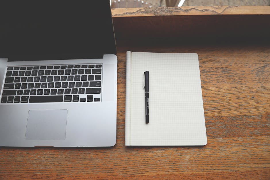 Minimalist workspace featuring a laptop, notebook, and pen on a rustic wooden desk.