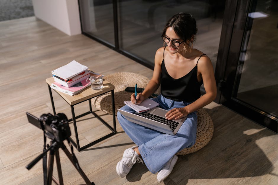 Young woman studying indoors with a laptop and camera setup, symbolizing remote learning.
