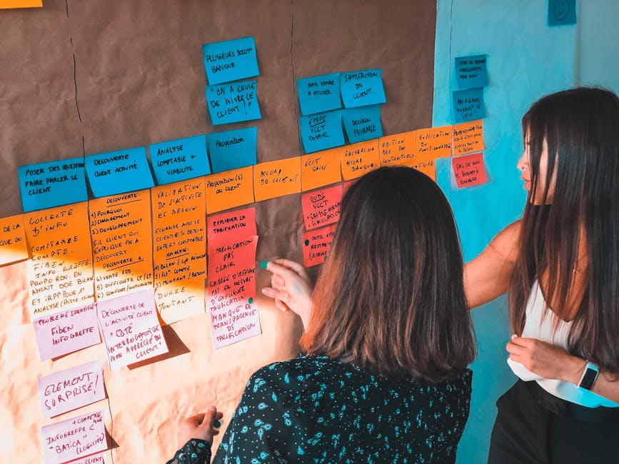 Two women engaged in collaborative planning using colorful Post-it notes on a board in a modern office space.