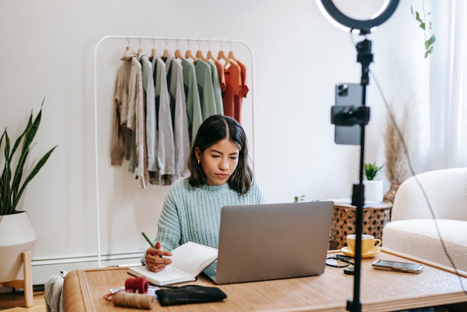 Young female freelancer sitting in light modern room while taking notes and using laptop in front of ring lamp and phone camera