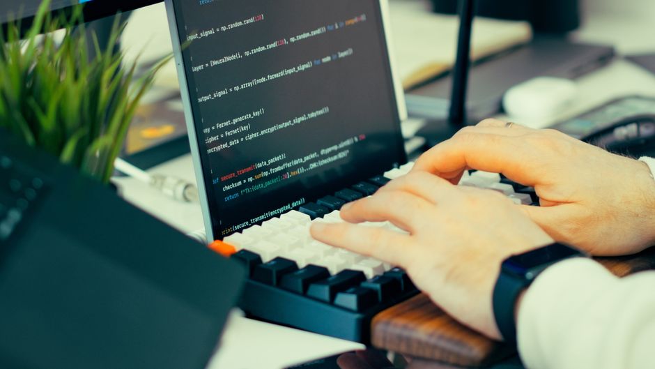 Close-up view of a developer typing code on a keyboard with a computer screen showing scripts.
