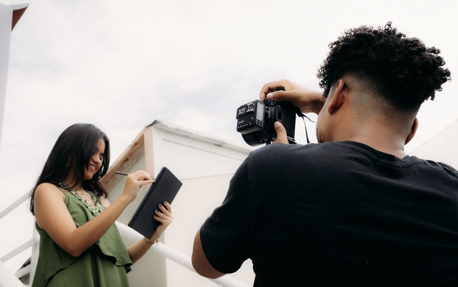 Photographer taking a picture of a smiling young woman holding a notebook outdoors.