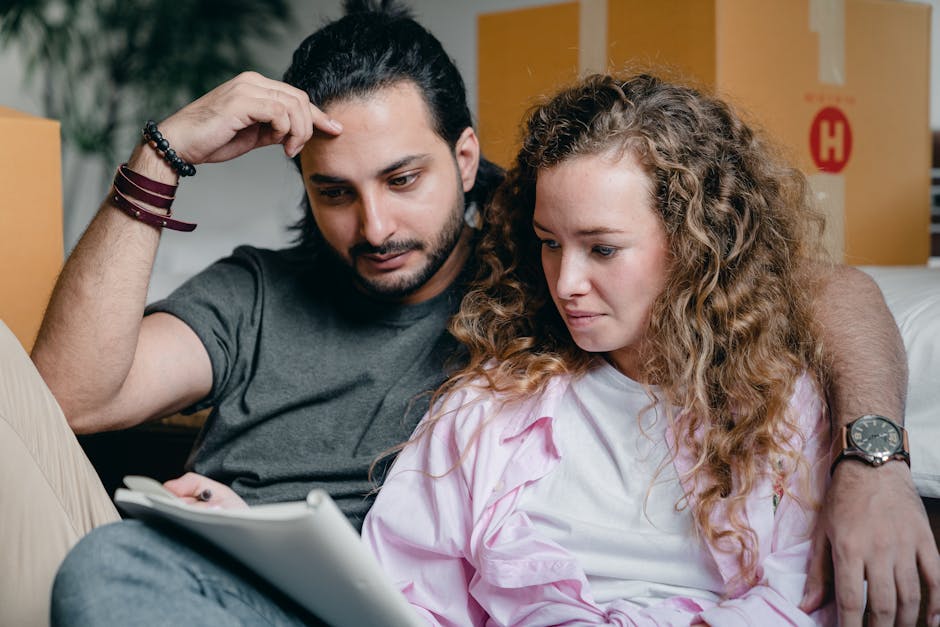Focused male and female in casual wear sitting near boxes and looking through notebook thoughtfully while moving house