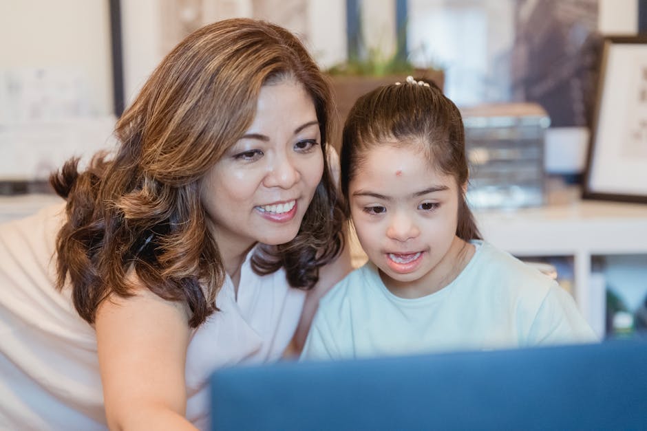 A mother and daughter with Down syndrome smiling while looking at a laptop indoors.
