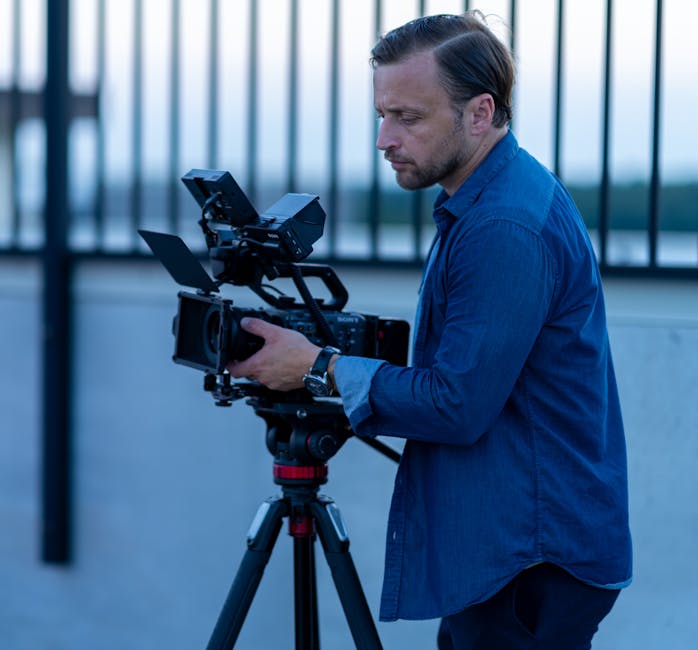 A male filmmaker in Chicago adjusting a video camera on a tripod, outdoors at twilight.